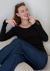 Woman sitting on a couch wearing a black top and blue jeans, holding a phone.