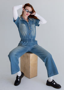 Person wearing a blue denim outfit sitting on a wooden block against a plain background