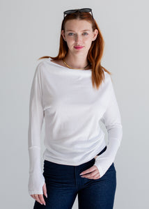 Woman wearing a white long-sleeve top and dark jeans against a plain background