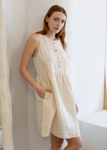Woman wearing a beige sleeveless dress standing against a white wall.