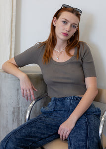 Woman sitting on a chair wearing a dark grey top and blue jeans, with a neutral background.