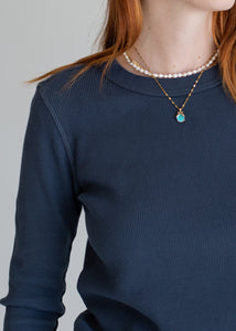 Person wearing a navy blue long-sleeve shirt with a pearl necklace and a gold necklace with a green pendant.
