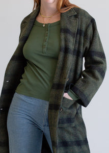 Person wearing a green plaid coat over a green shirt and blue jeans on a plain background