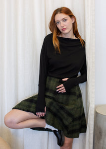Woman wearing a black top and green plaid skirt standing against a white curtain.