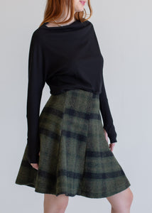 Person wearing a black long-sleeve top and green plaid skirt against a plain background
