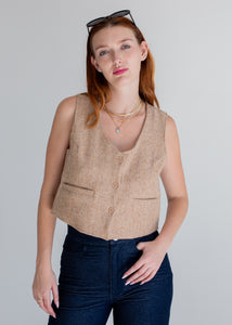 Woman wearing a beige vest with sunglasses on her head against a gray background.